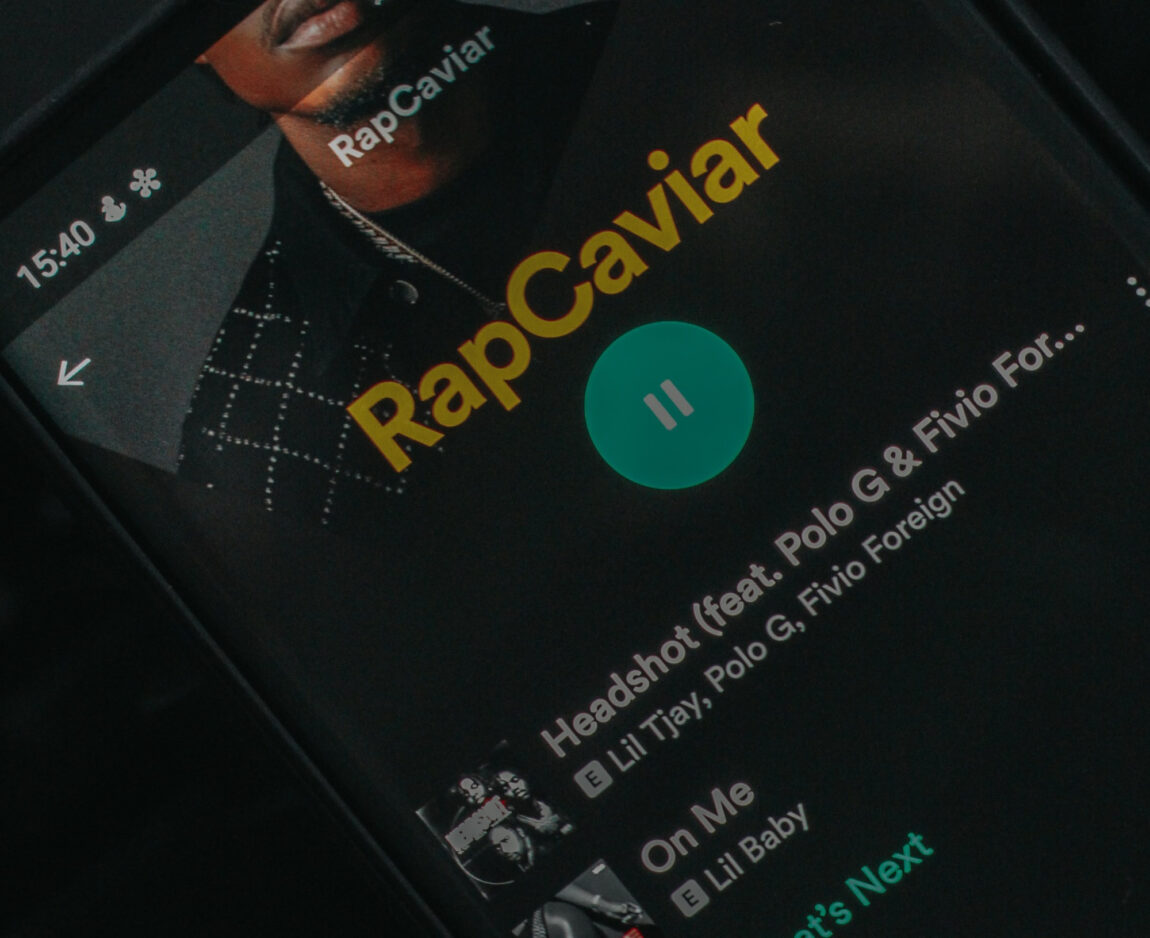 Close-up of a smartphone screen showing Spotify's RapCaviar playlist with the song 'Headshot' by Lil Tjay, Polo G, and Fivio Foreign currently playing.
