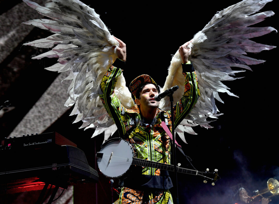 Sufjan Stevens on stage wearing a tracksuit and angel wings. His arms are extended upwards as he sings.