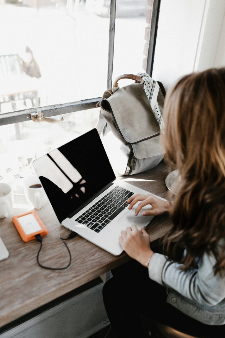 A woman works on a laptop at a wooden desk by a window. A gray backpack, orange hard drive, and coffee cup are nearby. Bright lighting.