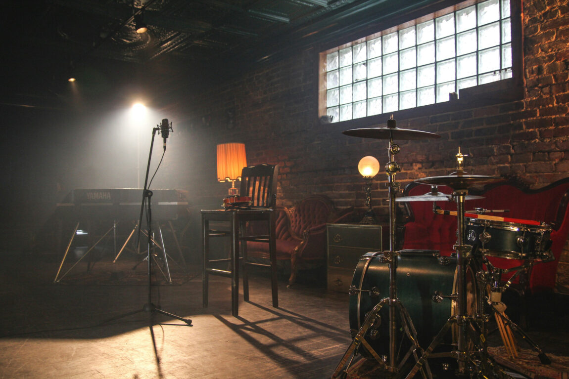 Dimly lit venue with drum set, microphone, keyboard, and vintage furniture. Warm light from a lamp creates a cozy atmosphere.