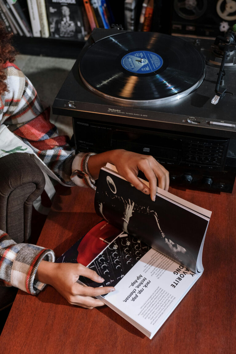 Person in plaid shirt flips pages of a magazine near a vintage record player. Books and albums visible in the background, cozy setting.