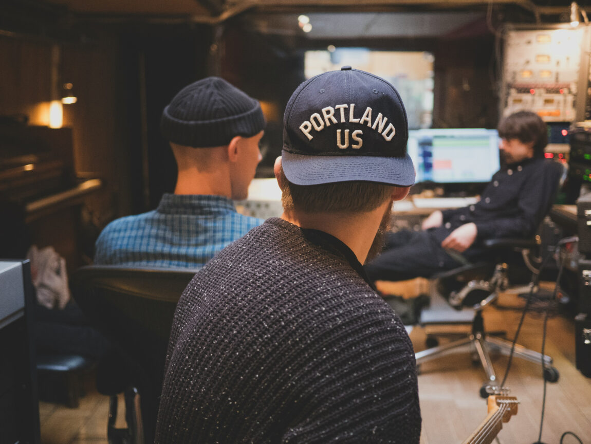 Three people in a recording studio. One wears a hat reading