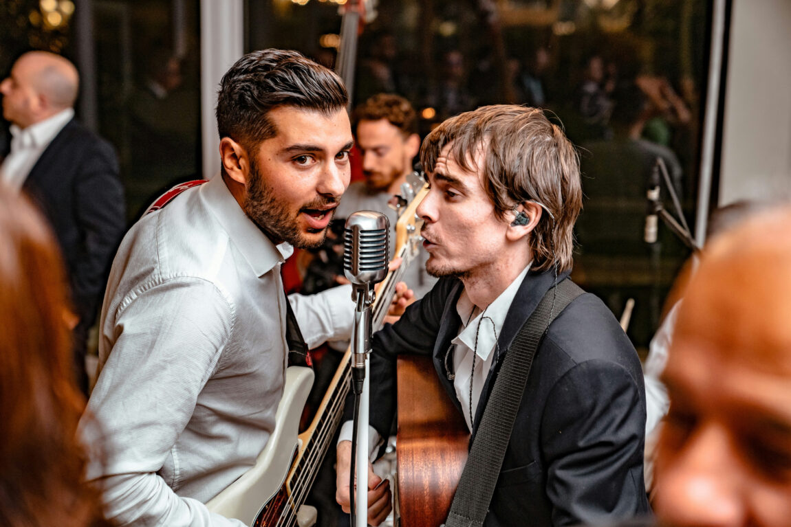 Two musicians sing passionately into a vintage microphone. One holds a guitar, the other holds a bass. Background is blurred with warm lighting creating an energetic vibe.