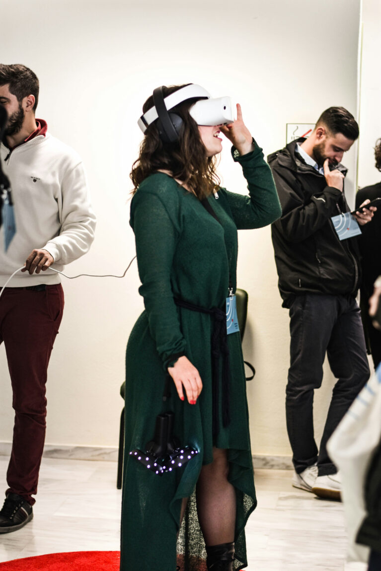 Woman in a green dress using a VR headset; background shows two men with phones. Bright indoor setting, tech event vibe.