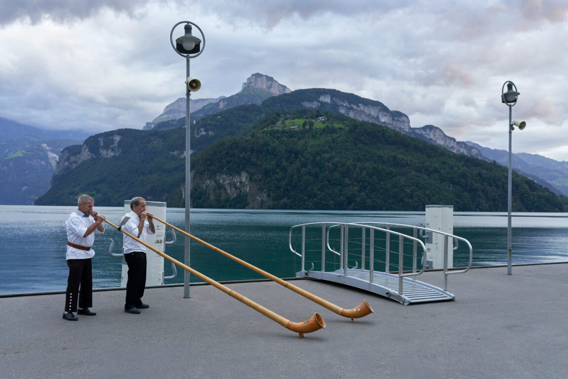 Two men play long alphorns on a lakeside pier, surrounded by mountains under a cloudy sky. The mood is serene.