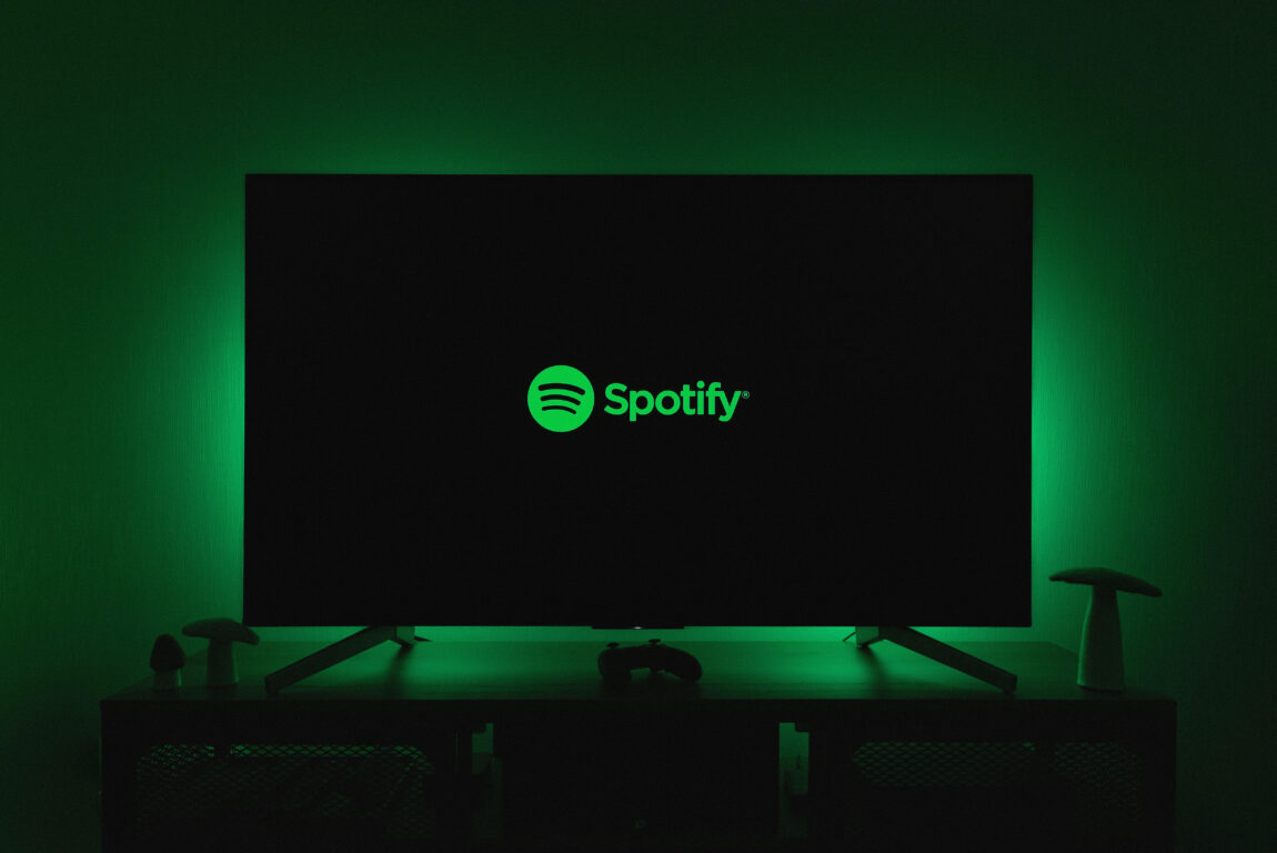 TV screen displaying the Spotify logo in green, set against a dark background with green ambient lighting. Mood is calm and focused.