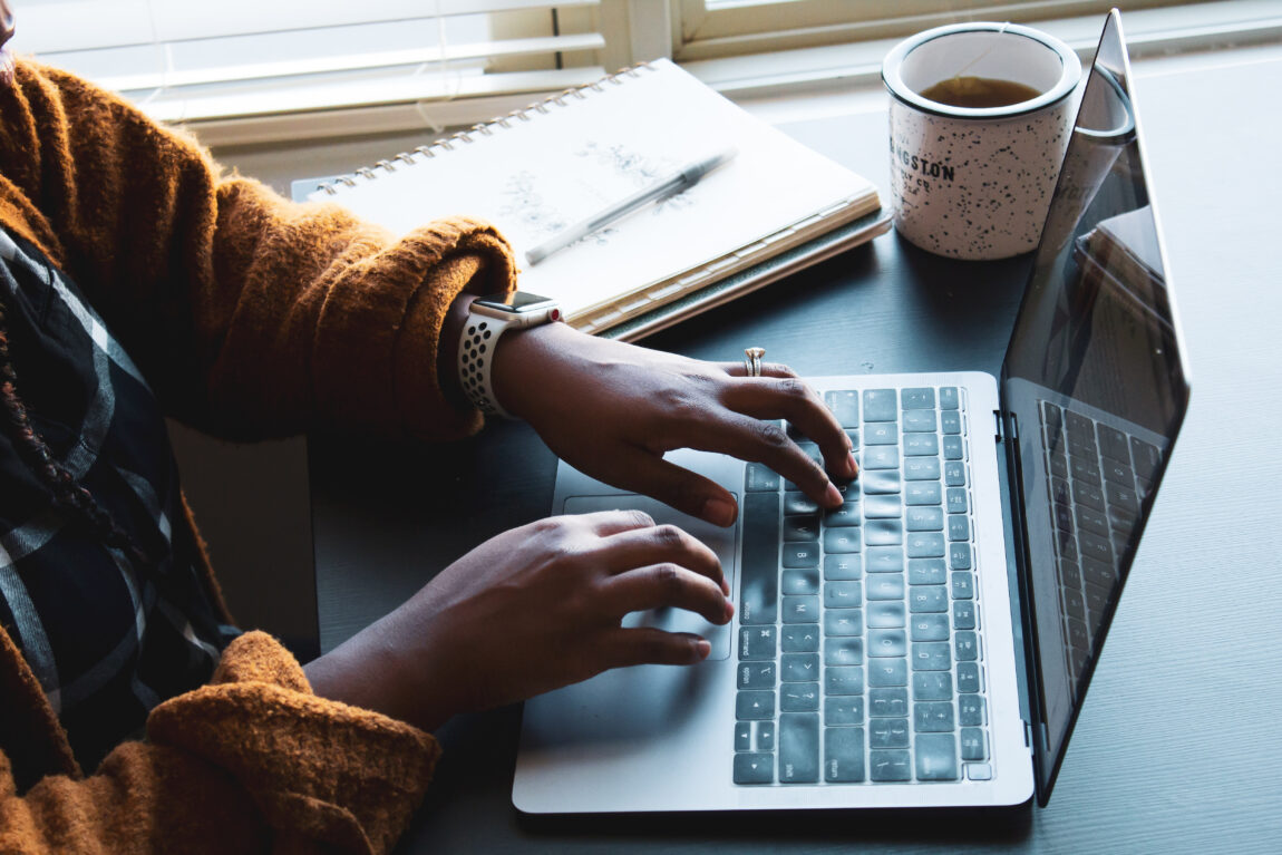Person wearing a brown sweater typing on a laptop, with a notebook and a mug of coffee placed on a desk near a window.