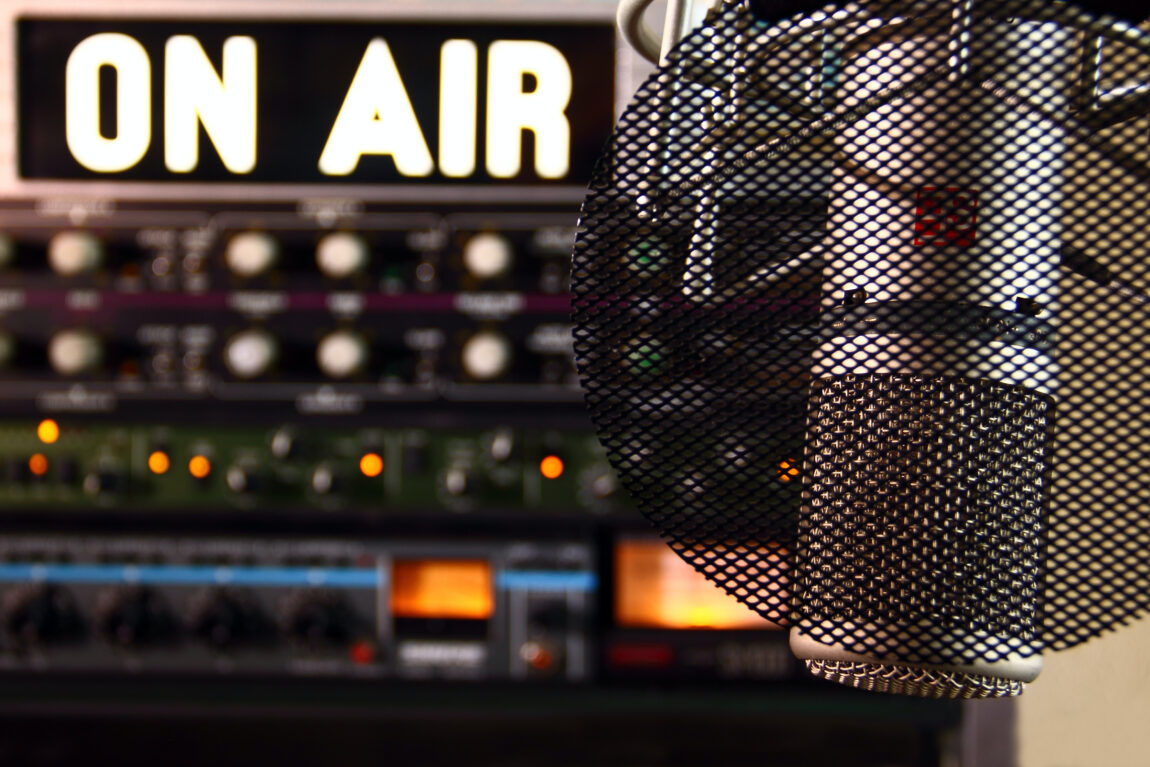 Close-up of a professional microphone with a pop filter in front of a glowing 'ON AIR' sign and audio equipment in a radio studio.